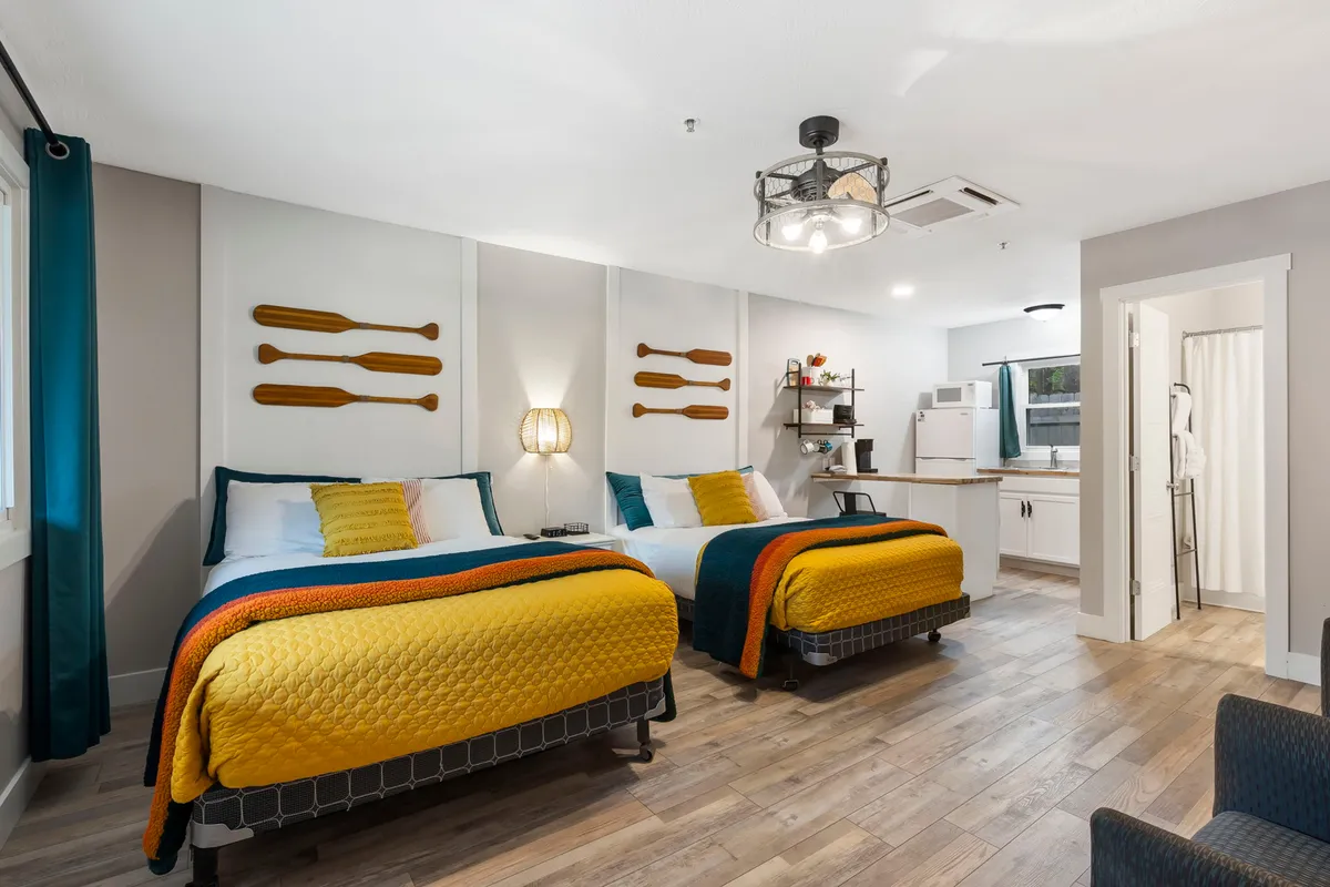 This image shows a cheerful and comfortable interior unit with two beds featuring vibrant bedding, wooden paddles mounted on the wall, a modern light fixture, and an open kitchenette area. The space is designed for relaxation, unwinding, and light meal preparation, making it perfect for families or small groups.