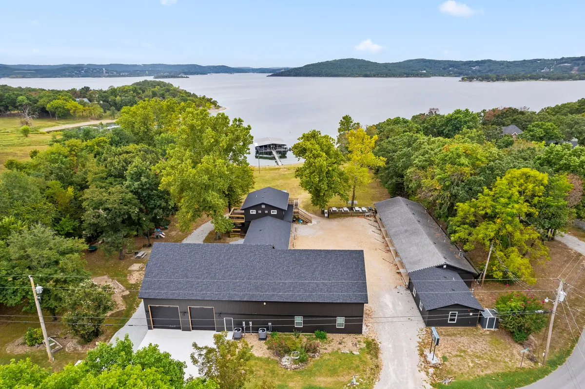 The image showcases a spacious lakefront home surrounded by lush trees, with an inviting private dock extending over the calm waters of Table Rock Lake. The sun is setting, casting a golden glow across the landscape, and open green spaces and a welcoming porch hint at opportunities for gathering, relaxation, and lakeside fun.