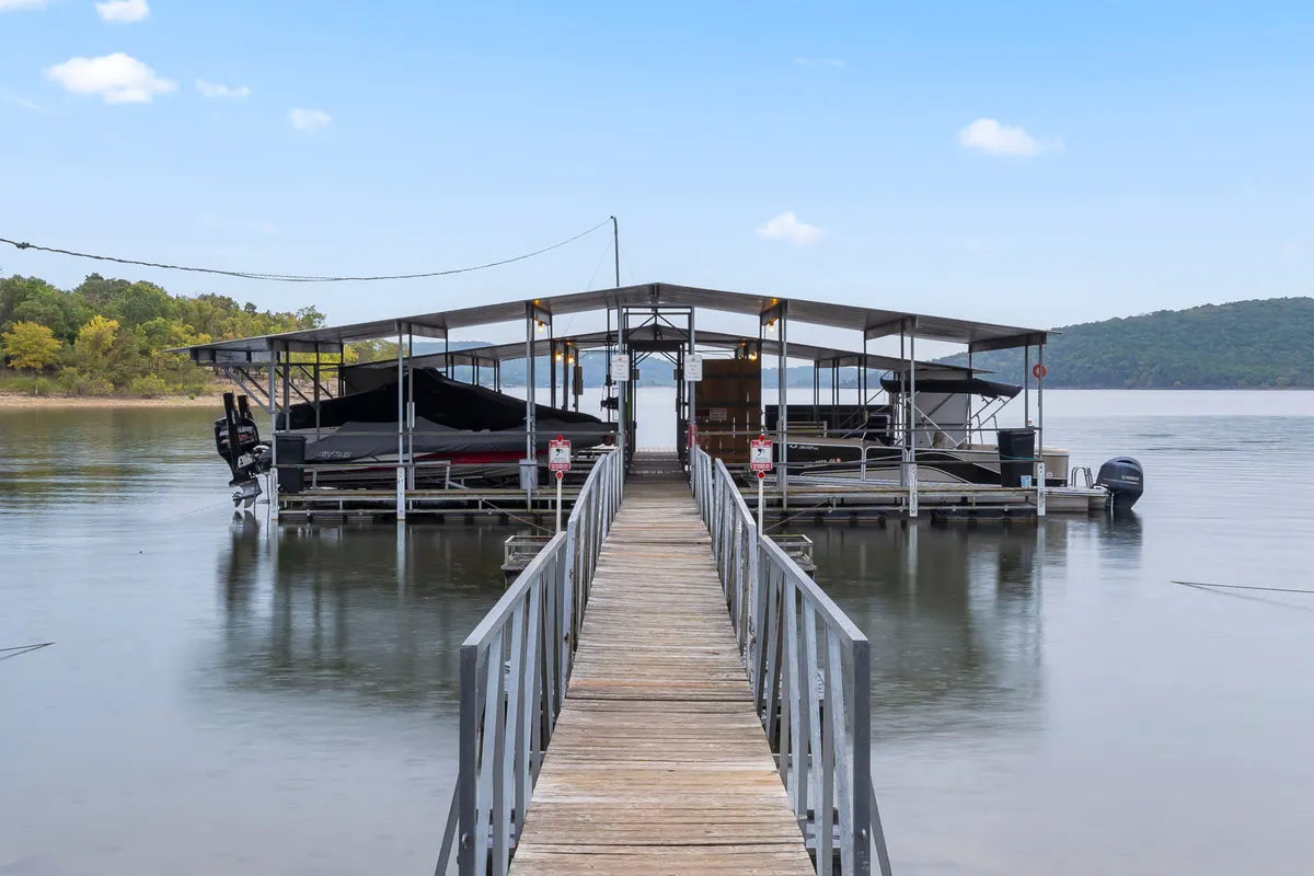 A tranquil lakeside scene featuring a dock extending over the water with boats moored and gentle hills in the background. The area appears perfect for boating, fishing, or a relaxing day by sun-dappled waters.