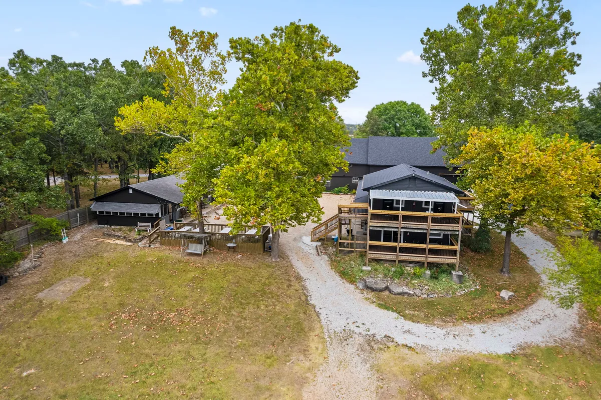 A wide exterior view of the main house framed by large trees, a deck area, and a gravel path. The scene conveys spacious grounds where multiple families or groups of friends could spread out and enjoy outdoor activities together.