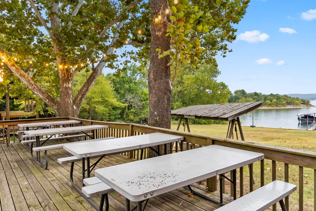 An inviting lakeside gathering space with wooden picnic tables shaded by large trees and twinkling decorative lights, all overlooking the shimmering lake. The setting is ideal for group meals, barbecues, and evening socializing, highlighting the communal spirit of the resort.